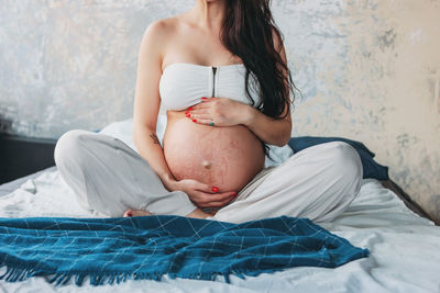 Low section of pregnant woman sitting on bed at home