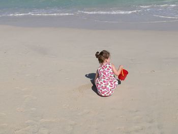 Rear view of girl holding toy bucket while crouching at beach