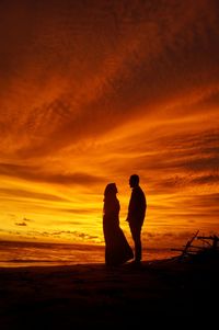 Side view of silhouette couple standing at beach against orange sky during sunset