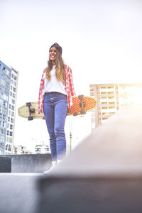 Beautiful young woman in cap is sitting at skatepark on the ramp with her longboard.