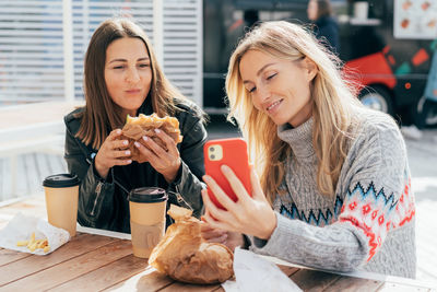 Women eat street fast food and chatting while sitting at a table outside and using phone for video.