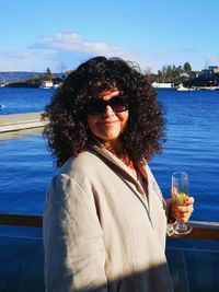 Portrait of woman with curly hair wearing sunglasses against sea in city