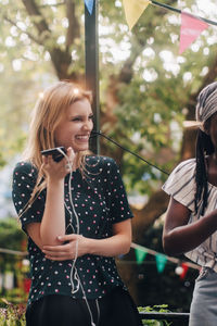 Smiling young woman holding mobile phone while standing by female friend in balcony during party