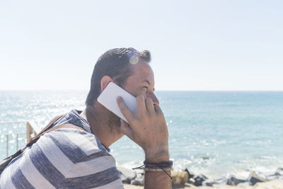 Side view of man listening over phone while standing at beach against clear sky