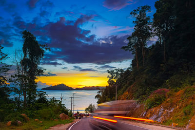 Road by trees against sky during sunset