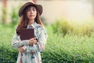 Beautiful woman looking away while holding book against plants