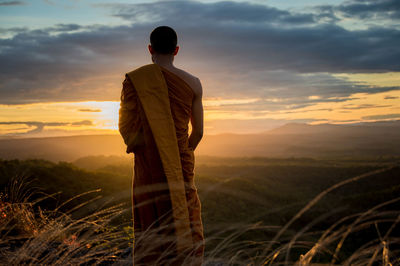 Rear view of monk standing on field against sky at sunset