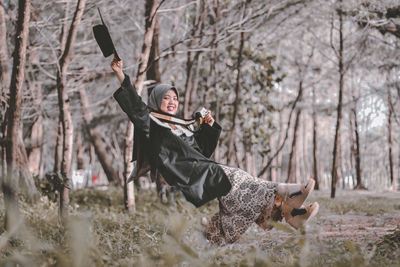 Full length of woman holding hat levitating against trees