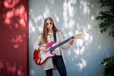 Portrait of young woman playing guitar while standing by wall