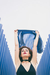 Portrait of young woman holding blue scarf amidst corrugated irons against clear sky
