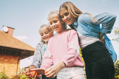 Low angle view of girls using smart phone standing against sky