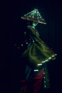 Anonymous female in authentic outfit and vietnamese hat with glowing illumination standing in dark modern studio on black background during performance