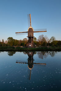 Traditional windmill by lake against clear blue sky