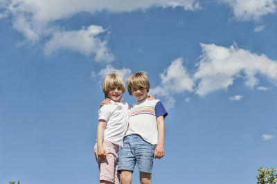 Low angle of adorable brothers hugging against blue sky in summer while looking at camera and enjoying weekend
