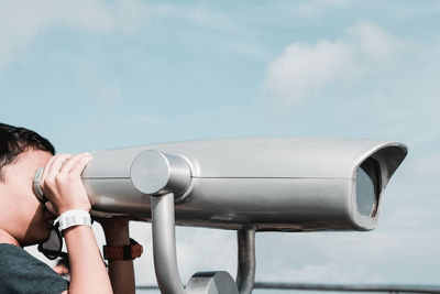 Side view of boy looking through coin-operated binoculars against sky