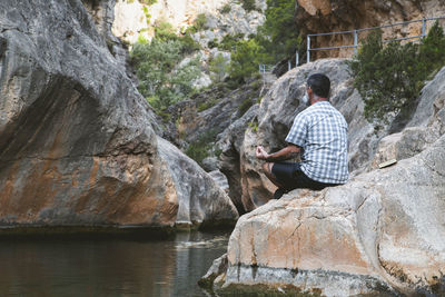 Man from behind meditating on top of a rock at the foot of a mountain river