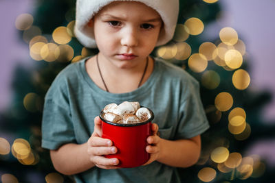 Little boy in santa hat holding a cup of hot chocolate with marshmallow