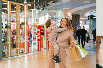 Smiling mother carries her little happy son in her arms, holds shopping bags in her hands