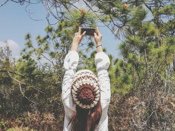 Rear view of woman photographing on mobile phone against trees