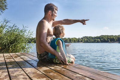 Father pointing while sitting with son on wet pier over lake against sky