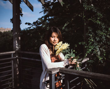Young woman standing with flowers by railing against trees