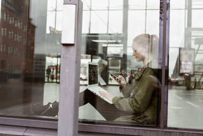 Woman using laptop and smart phone while sitting by window at station