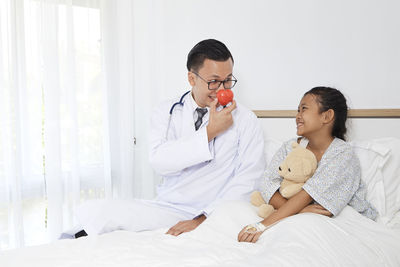 Playful doctor sitting with girl on bed at hospital