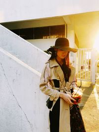 Young woman holding camera while standing against built structure