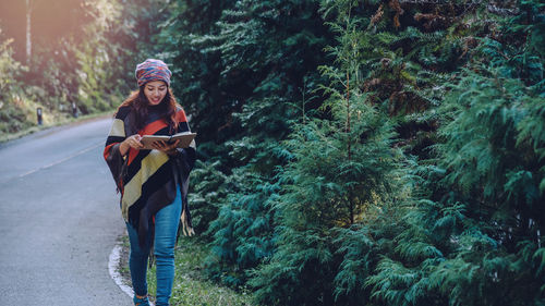 Full length of young woman reading book while walking by trees on road