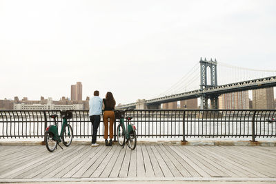 Couple standing with bicycles on footpath by manhattan bridge against sky