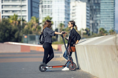 Woman talking with friend while standing with push scooter on road in city