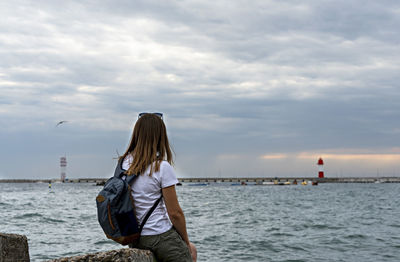 Rear view of young woman looking at view of sea with lighthouse and cloudy sky at sunset, landscape