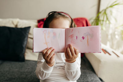 Unrecognizable little girl demonstrating sketchbook with doodles while sitting on sofa and drawing at home