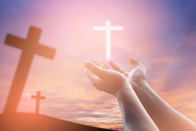 Low angle view of hands gesturing towards cross at sunset
