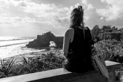 Rear view of woman sitting on bench while looking at batu bolong temple in sea against sky