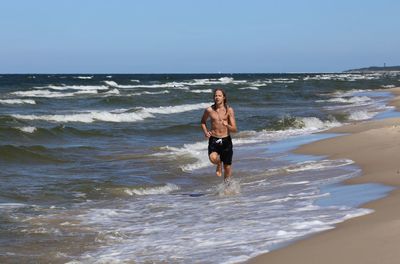 Shirtless teenager running on shore at beach against sky