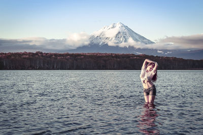 Young woman with arms raised standing in lake against sky during sunset