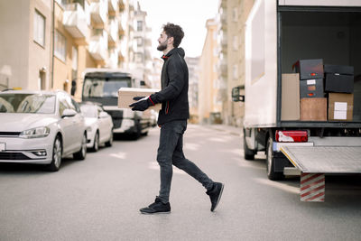 Side view of young delivery man carrying box while walking on street in city