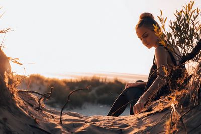 Side view of young woman sitting on tree trunk at beach against clear sky during sunset