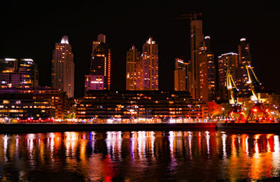 Illuminated buildings by river against sky at night