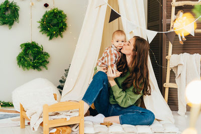 Young woman and baby playing, hugging and kissing while sitting in tent on christmas day at home