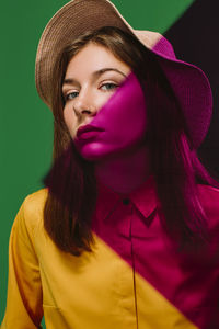 Young female model in stylish hat with red shadow on face and shoulder looking at camera against green background