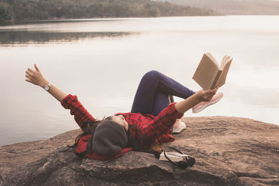 Young woman sitting on rock by lake against sky
