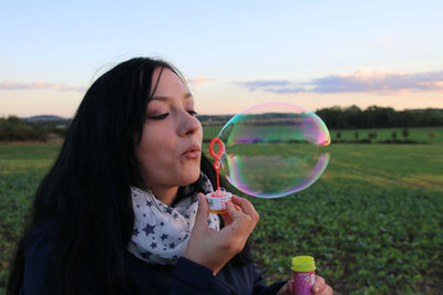 Side view of young woman blowing bubble from wand on grassy field