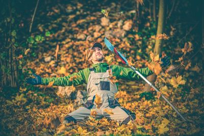 Mid adult man with arms outstretched kneeling on autumn leaves by gardening fork