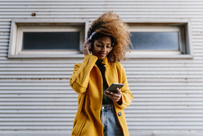 Smiling afro woman using mobile phone while listening music through headphones against wall