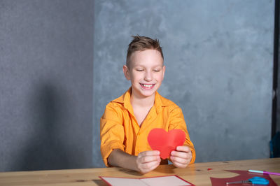 A cute boy is sitting at a table and holding a red heart in his hands for a valentine's day card
