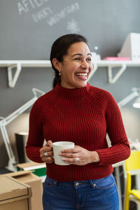 Cheerful businesswoman holding coffee cup while looking away against wall at creative office