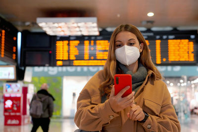 Female commuter checking her smartphone with behind timetables of departures arrivals.