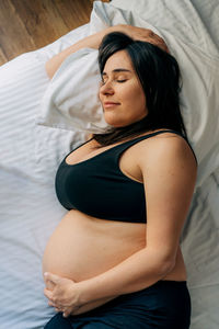 Middle-aged brunette pregnant woman hugging her tummy at home while lying on bed